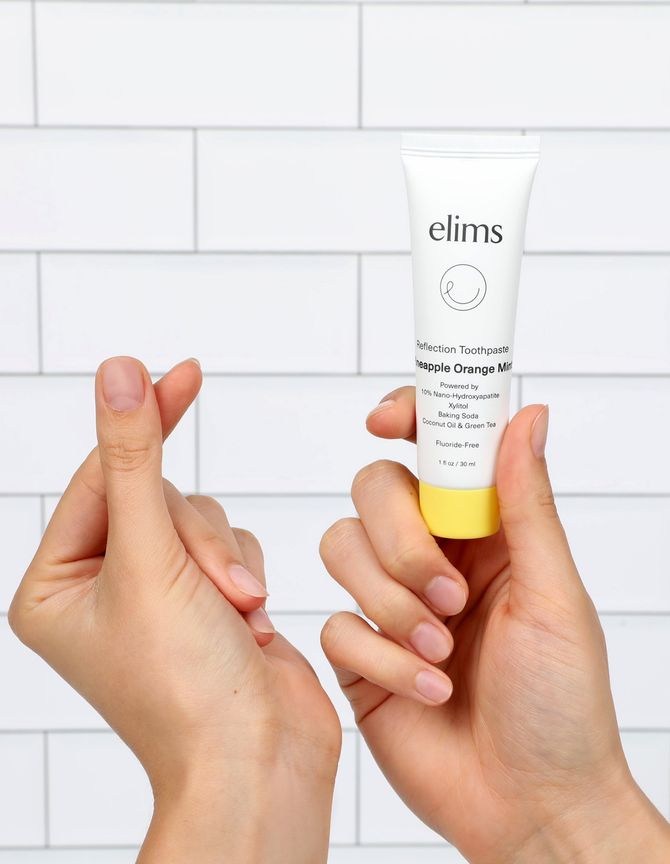 ELIMS Reflection Toothpaste in Pineapple Orange Mint flavor on a  a marble background with a hand holding a heart sign.